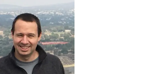 A smiling light-skinned man with short, dark hair and squinting eyes. He is wearing a black zip-up jacket over a blue-gray t-shirt. Behind and below him is a wooded area and a town with low mountains behind it.